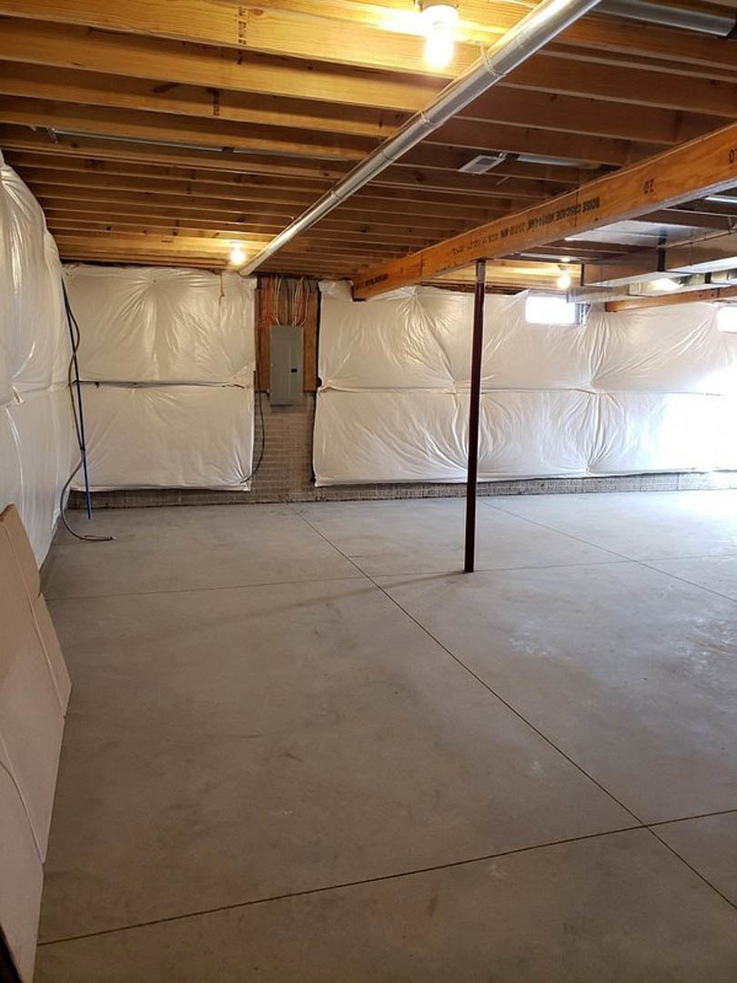 Open room with exposed wooden ceiling beam, white plaster wall partially covered by a plastic sheet, concrete floor, visible ceiling pipe, and minimal furnishings.