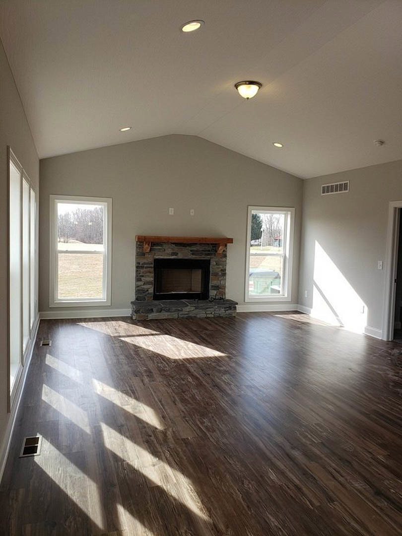 Spacious living room with wood flooring, stone fireplace featuring a wood mantel, large window with white frame overlooking yard