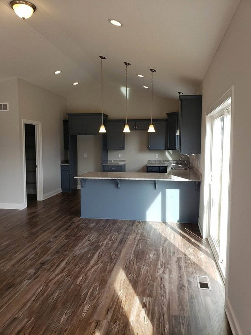 Open kitchen with marble-topped bar, wood plank flooring, white cabinetry, and pendant light fixture; sunlight streaming through a nearby white door.