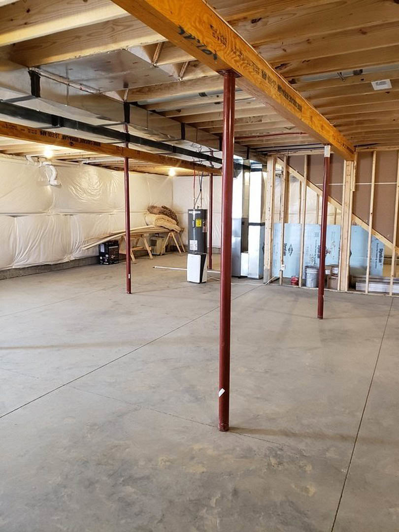 Open room with exposed wood ceiling beam, concrete floor supported by red steel poles, large white plastic wall covering, stack of blankets on a table, and blurry black box in the