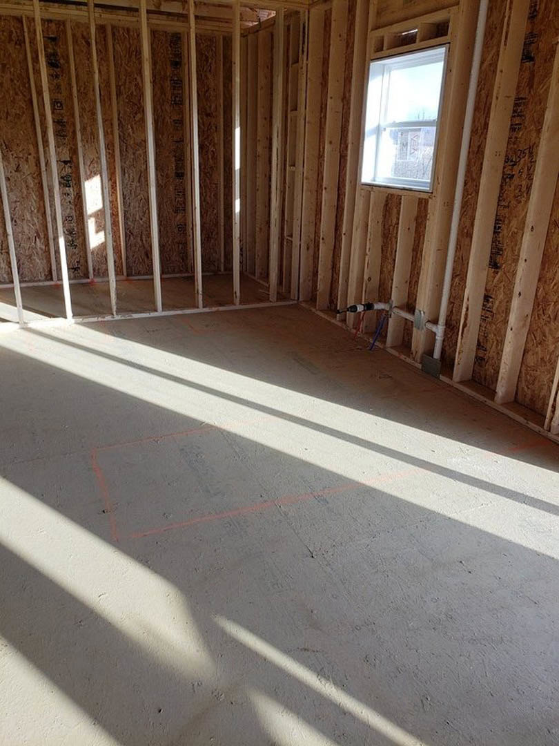 Unfinished room with exposed wood framing, concrete floor marked with red lines, two white-framed windows, and a close-up of a wood panel