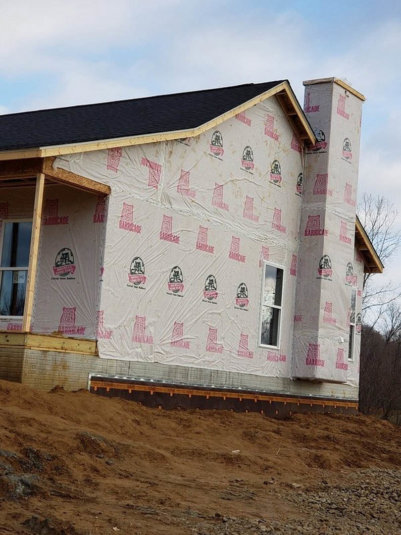 Two-story house under construction with white-framed windows, black roof, plastic sheeting covering exterior walls, and dirt hill in foreground