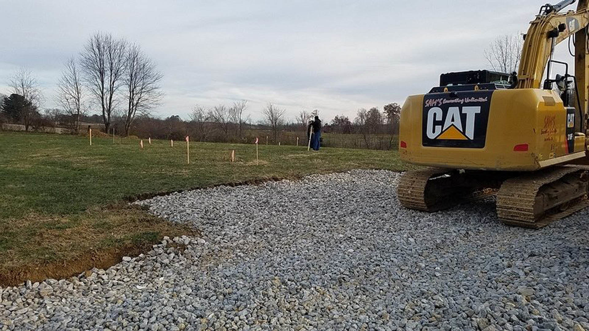 Yellow bulldozer parked on gravel driveway beside grassy yard, surrounded by trees under blue sky