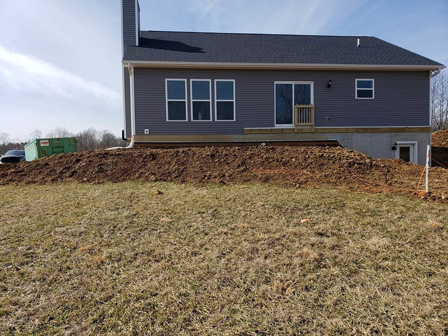 Two-story house with gray siding and white-framed windows, front porch, grassy yard with exposed dirt mound, wooden fence, green dumpster with signage, and train tracks in the