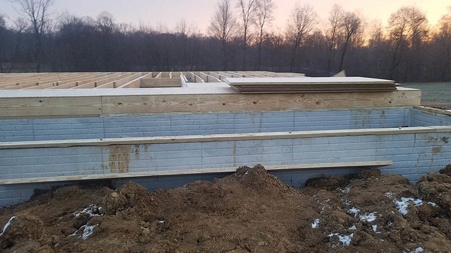 Wood-framed custom home under construction with exposed beams, unfinished walls, and a large dirt pile in the foreground; trees and open sky in the background
