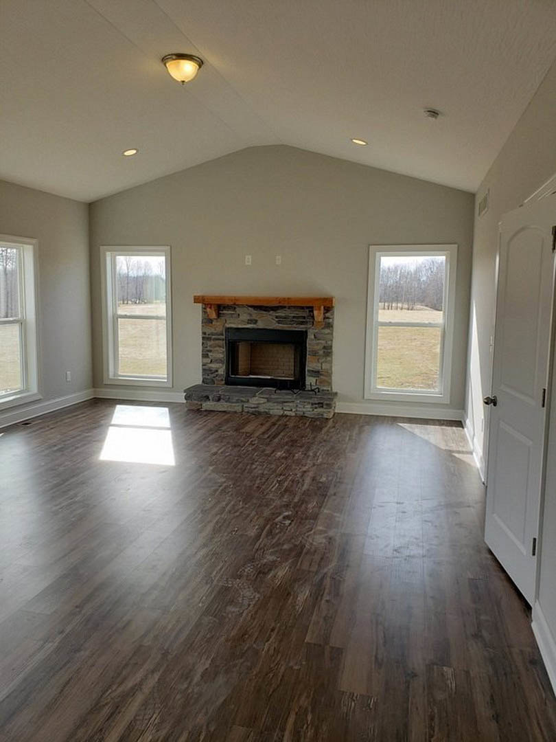 Living room with hardwood floors, stone fireplace featuring a wood mantel, large window overlooking a field and trees, white walls, and recessed ceiling lighting