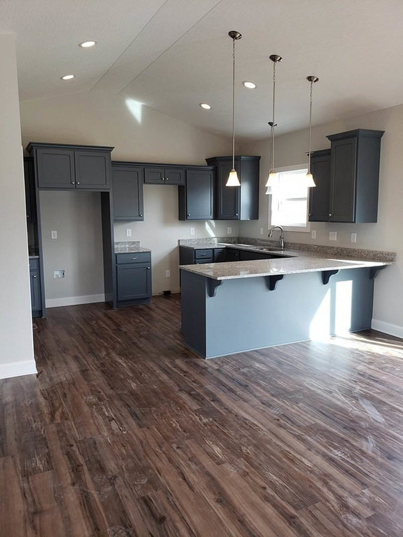 Kitchen with wood flooring, black cabinetry, stone countertop bar, stainless steel sink, and sunlight streaming through window onto floor and surfaces