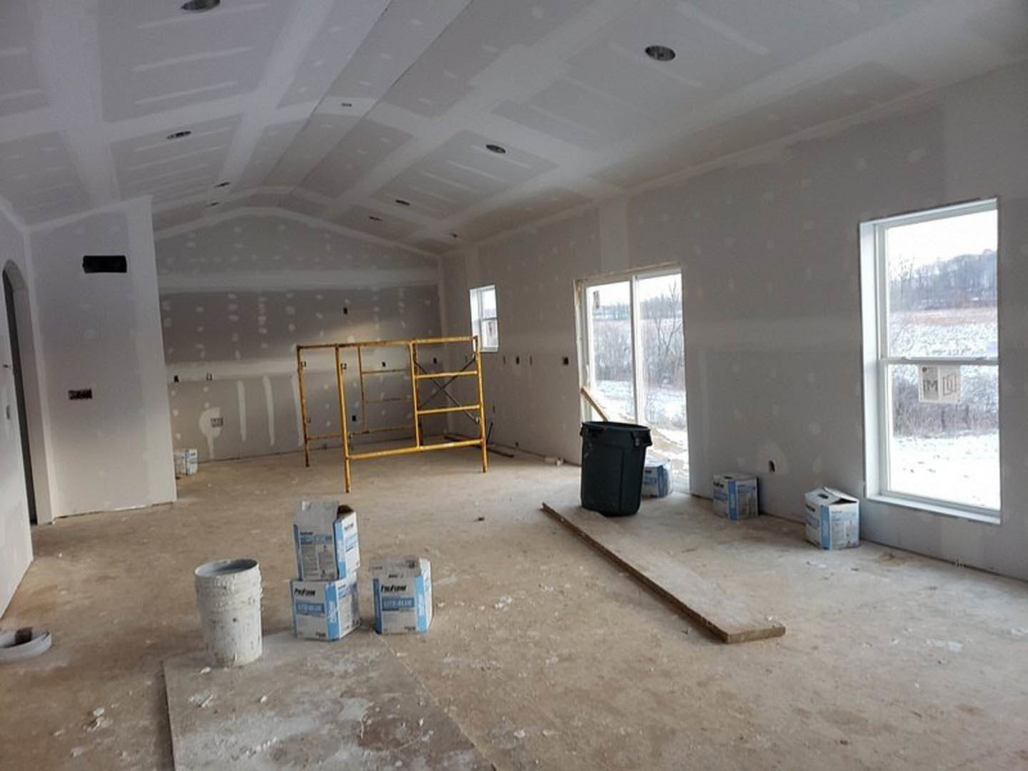 Unfinished room with white plaster walls, metal scaffolding, yellow ladder, black trash can, wood plank on floor, window displaying sign