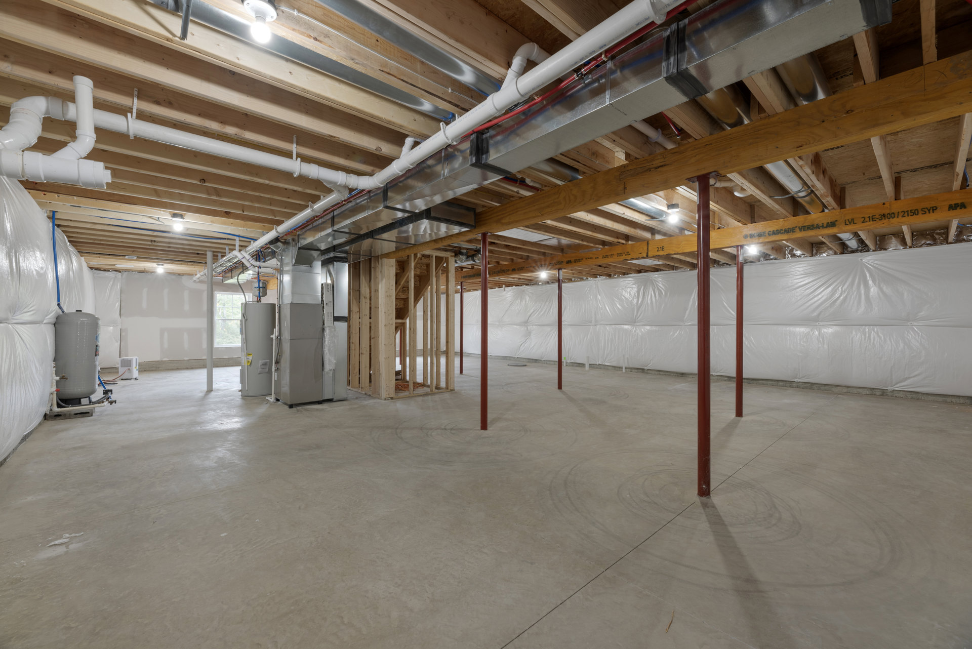 Unfinished basement with exposed ceiling pipes, concrete floor, red steel support pole, wooden framing, white water heater with yellow label