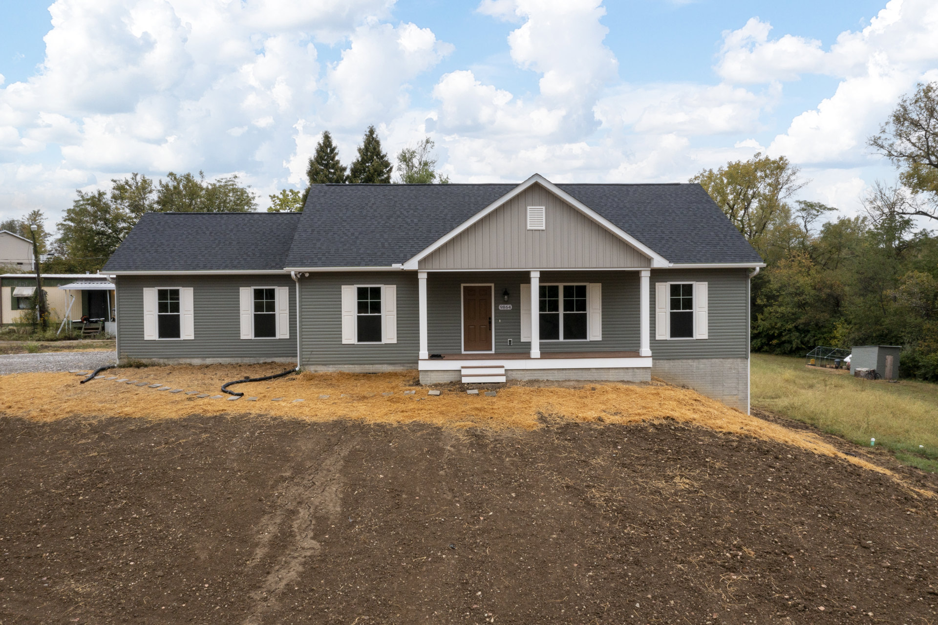White cottage with covered porch, front door, and windows, set behind a dirt hill with trees and clouds in the background