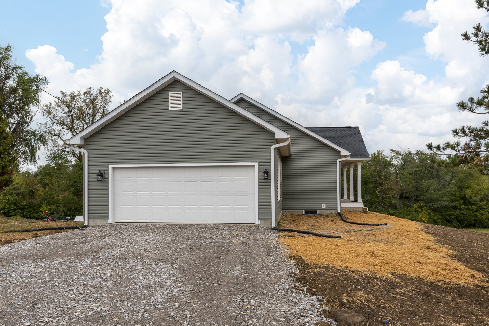 White garage door beneath gray roof, gravel and rocks in front, gray siding with white vent, dirt patch and black hose beside white house, cloudy sky and trees in background