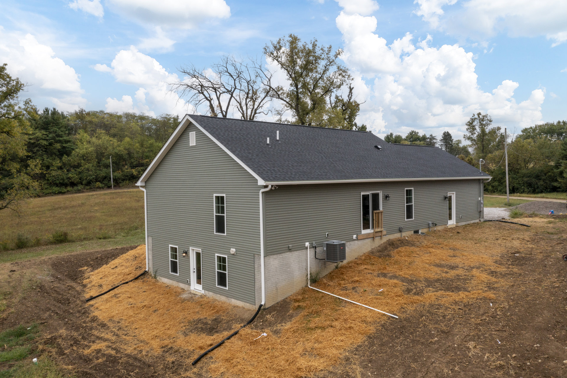 Partially built house with exposed framing, gray heat pump unit beside exterior wall, green lawn, garden hose on ground, wooden chair near doorway, mature trees in background.