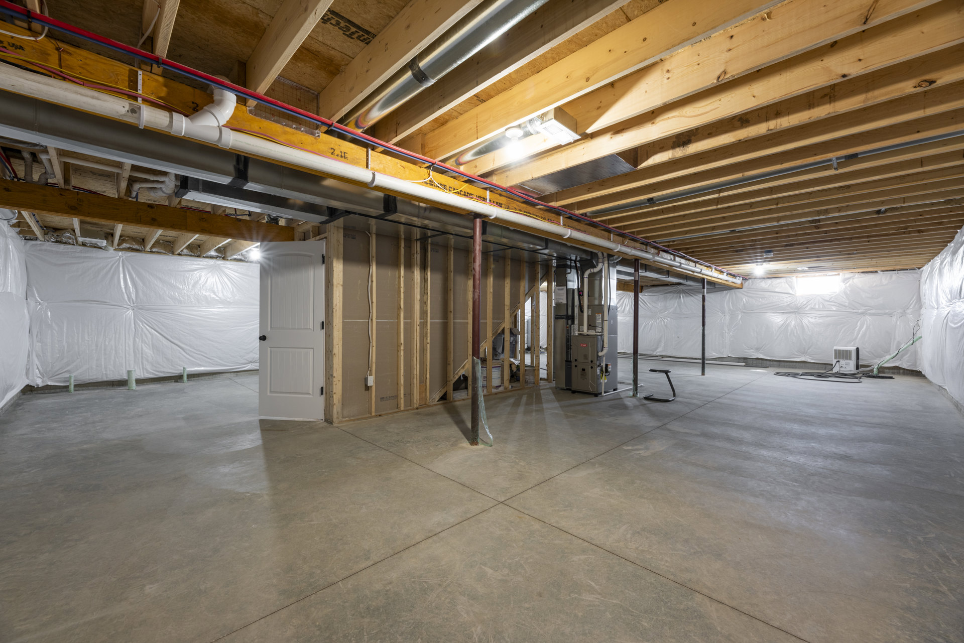 Basement room with exposed steel beam, concrete floor featuring a pipe, white door with black knob, white rectangular device with black wire, white plastic sheet covering part of