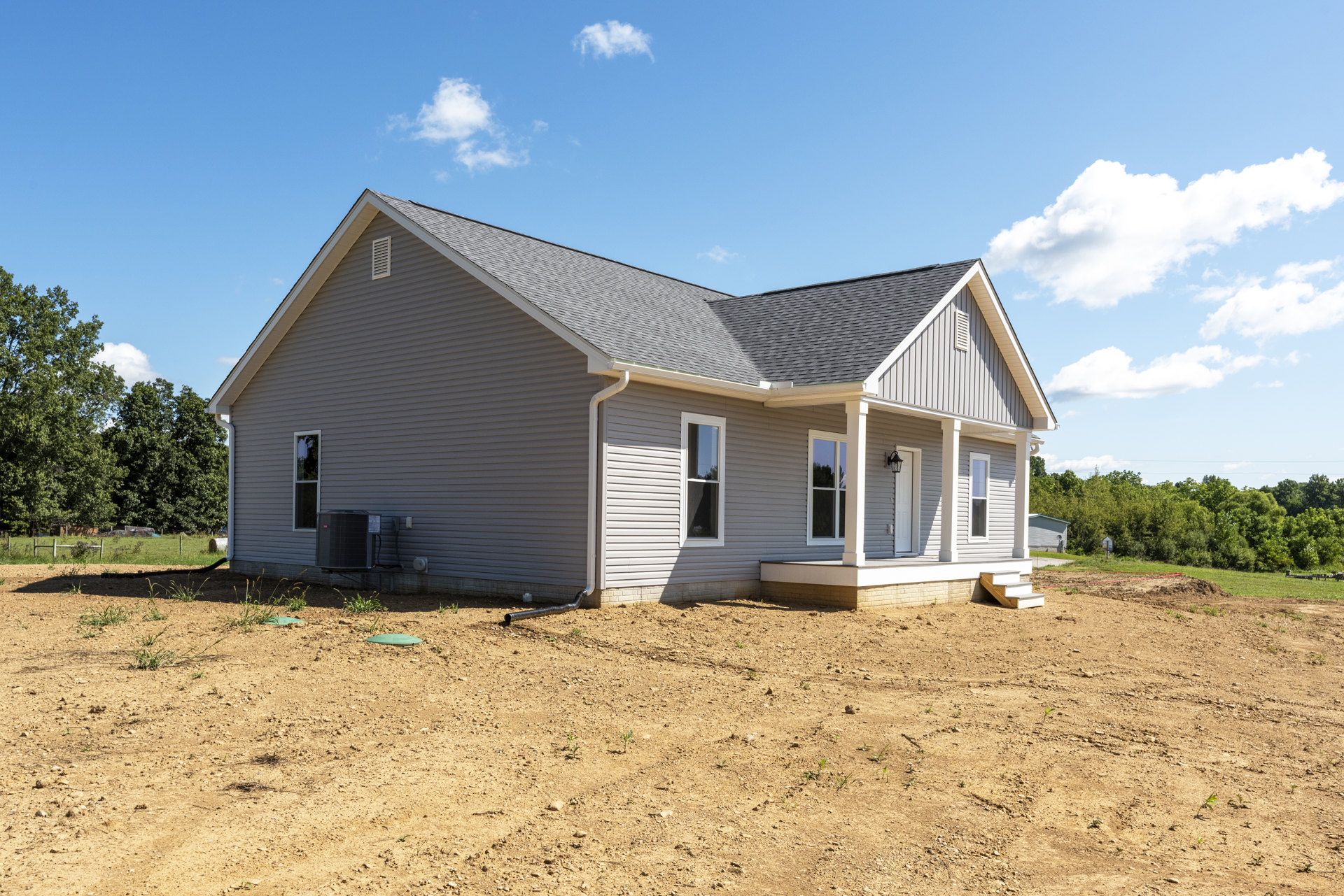 Modern house with white-framed windows reflecting trees, black heat pump on exterior wall, gutter along side, surrounded by dirt field under partly cloudy sky