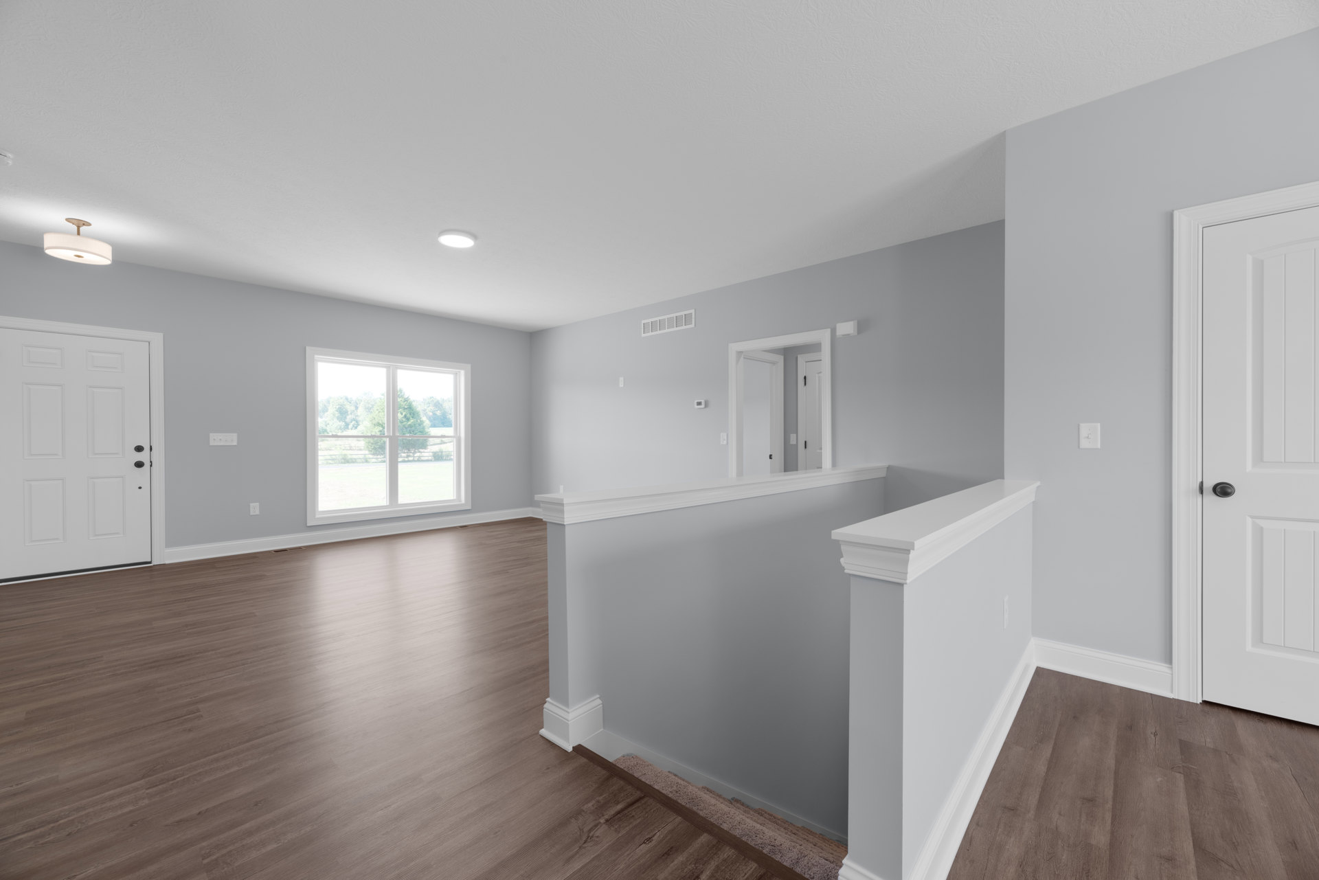 Wood floor room with white walls, white baseboards, a white door with black knobs, hallway leading to staircase, window showing trees outside