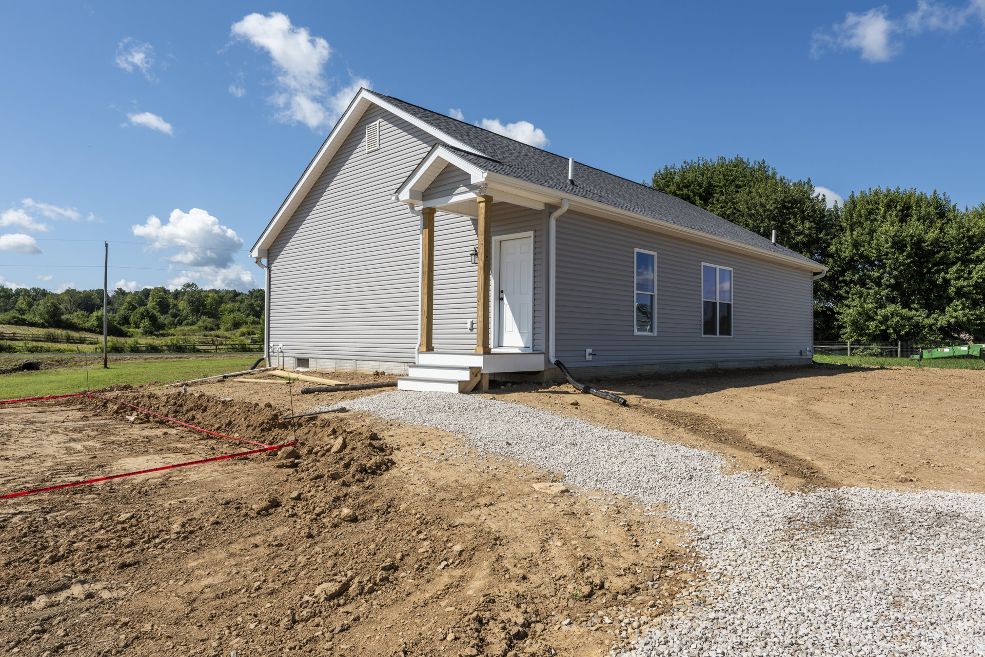 Two-story house under construction with exposed framing, gravel foundation, white front door with black handle, surrounded by dirt and mature trees