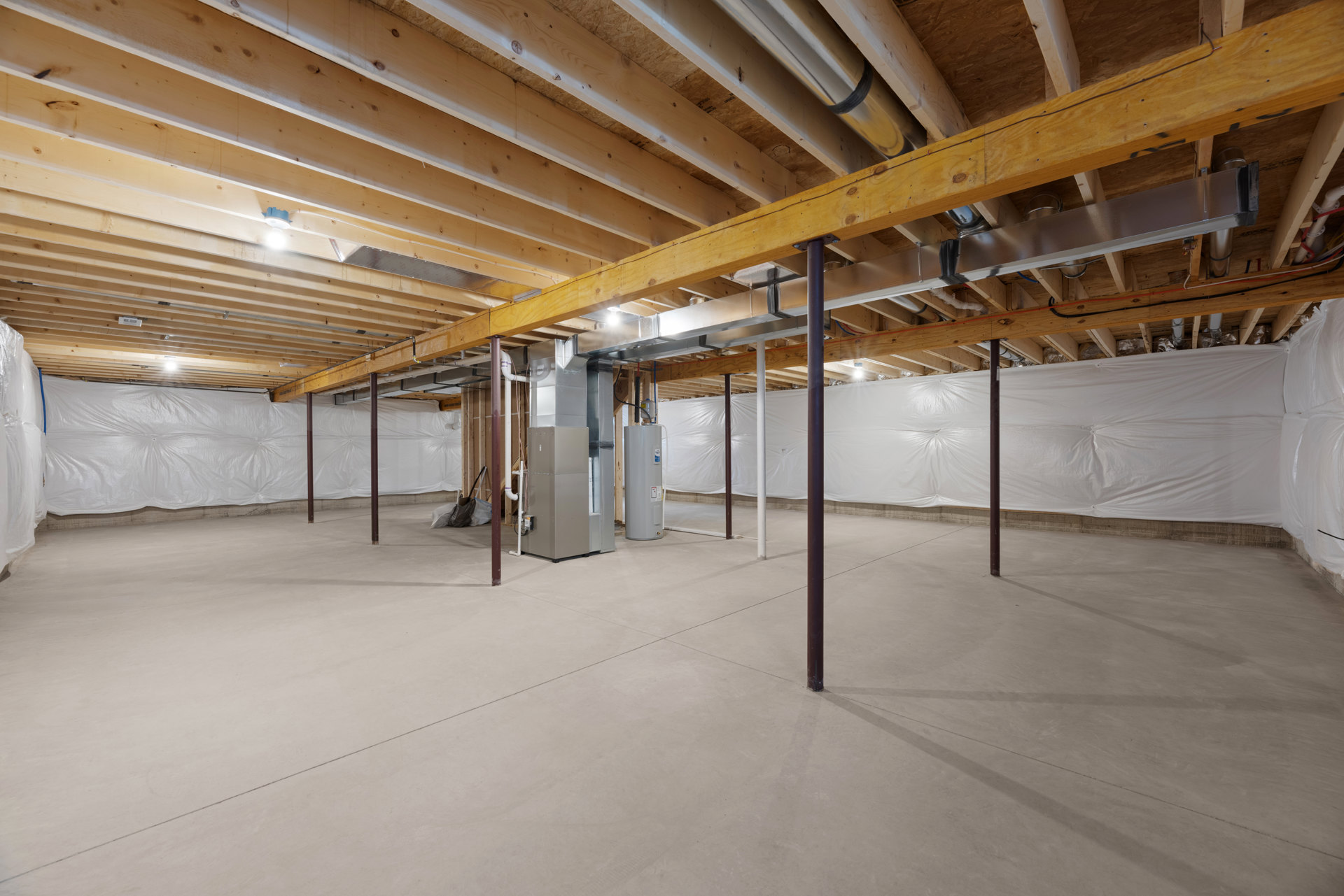 Open room with exposed wooden ceiling beams, polished concrete floor, stainless steel refrigerator, and visible pipes along unfinished walls.