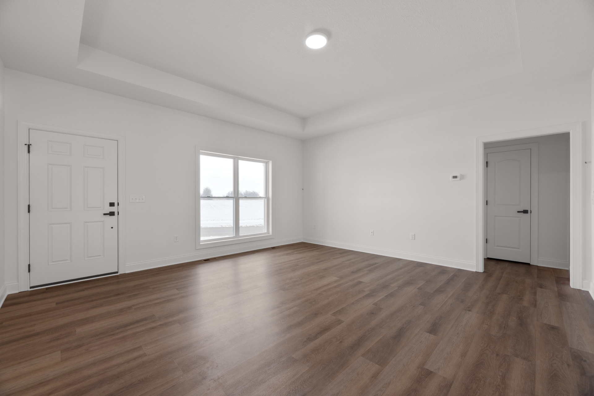 Room with hardwood floor, white door with black handles, ceiling light, and window overlooking snowy field