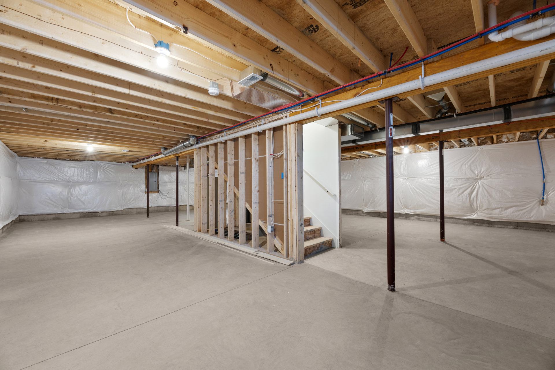 Open room with exposed wood beams, unfinished ceiling with visible pipes, white plastic wall covering, staircase with metal handrail, concrete floor.