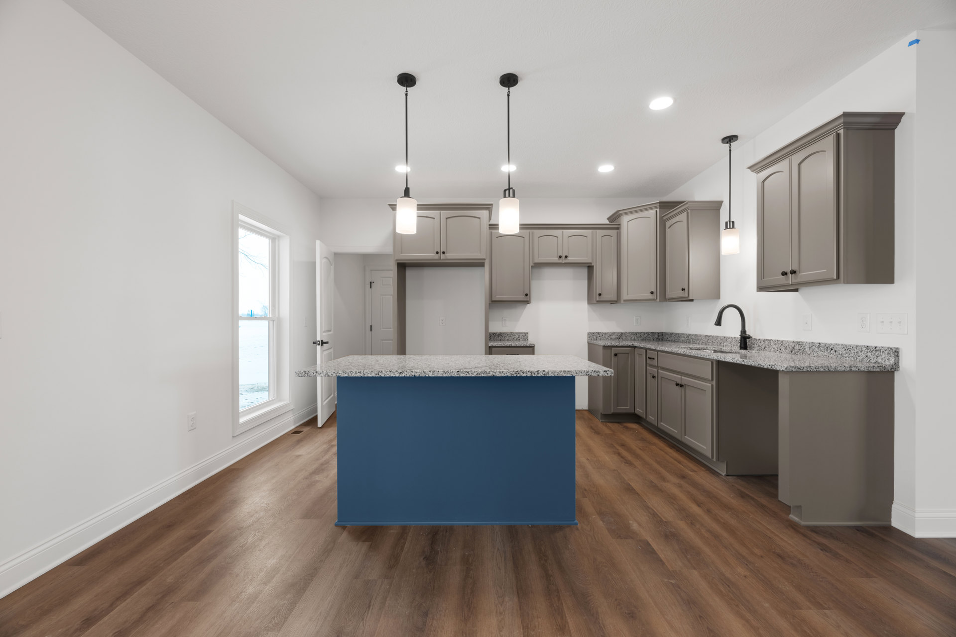 Blue kitchen island with white countertop, gray cabinets, stainless steel appliances, white door, large window overlooking snowy landscape, light wood flooring, and blue accent