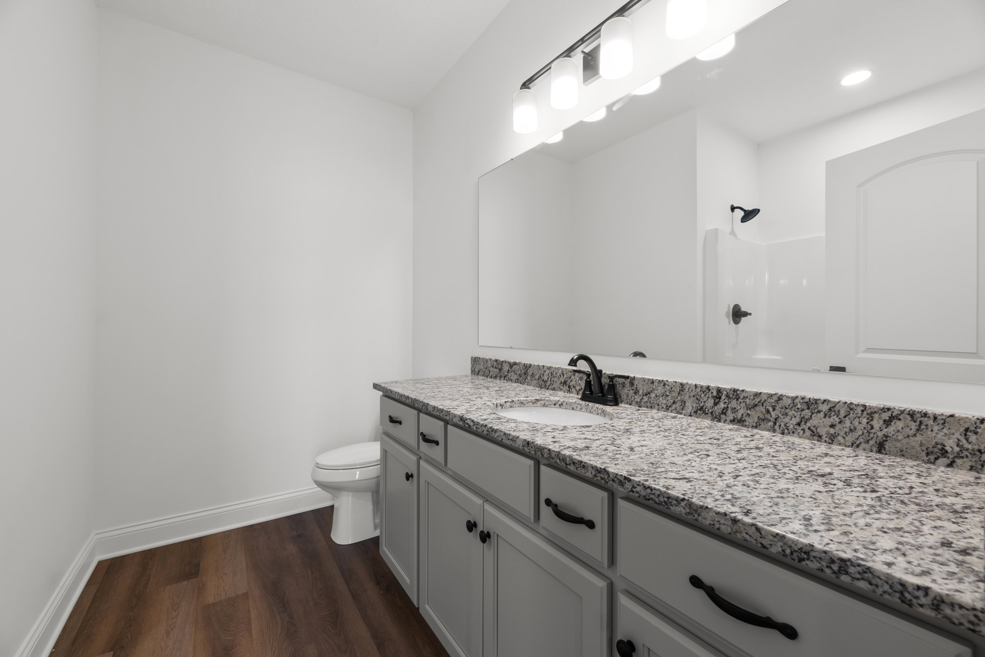 Bathroom with marble countertop, undermount sink, white drawers with black handles, wood flooring, white baseboard, curved-top white door, and toilet.