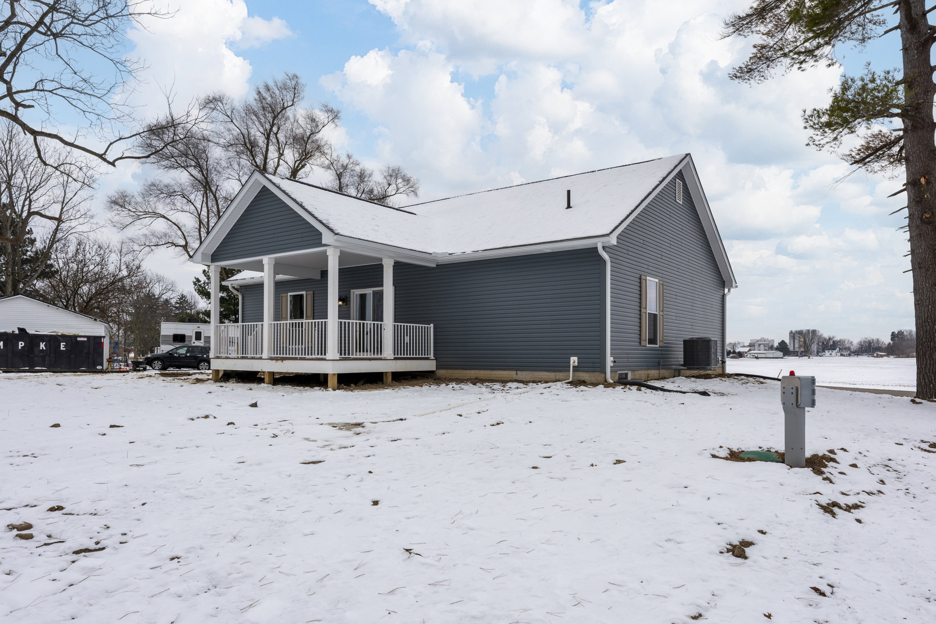 Two-story house with white siding, covered front porch, snow-covered yard, bare trees, and a black car parked beside a trailer.