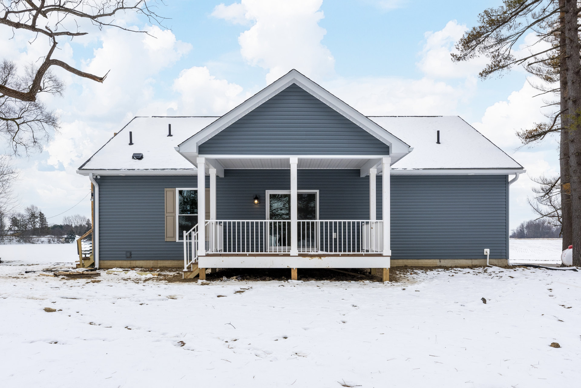 Front porch with white railing, snow-covered yard, light-colored siding, large windows, bare trees, overcast sky
