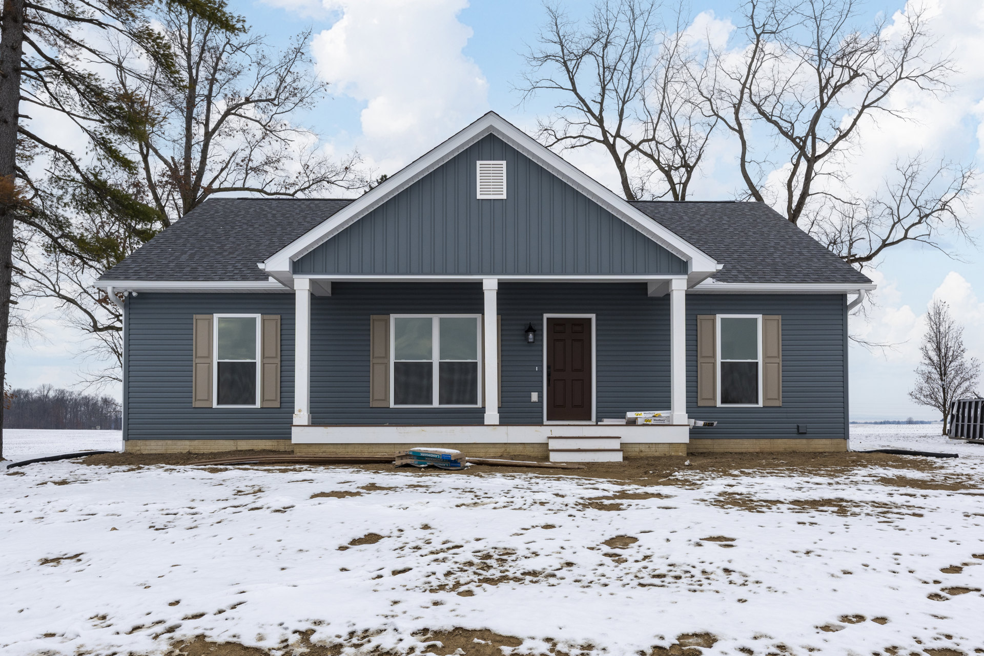 Two-story home with gray siding, white trim, and large windows; snow blankets the ground and porch, with piles of snow near the entry door and a screened window visible on the
