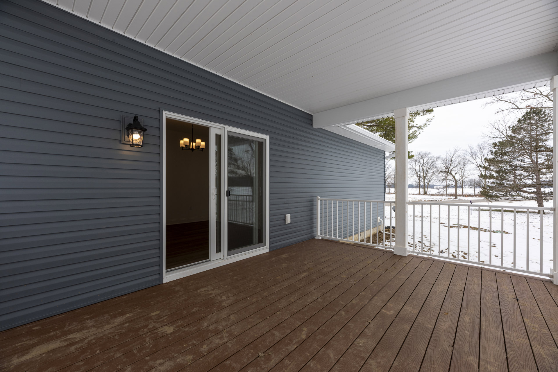 Wood deck with white railing, glass door featuring black handle, wall-mounted light fixture, chandelier visible inside, leafless trees in background