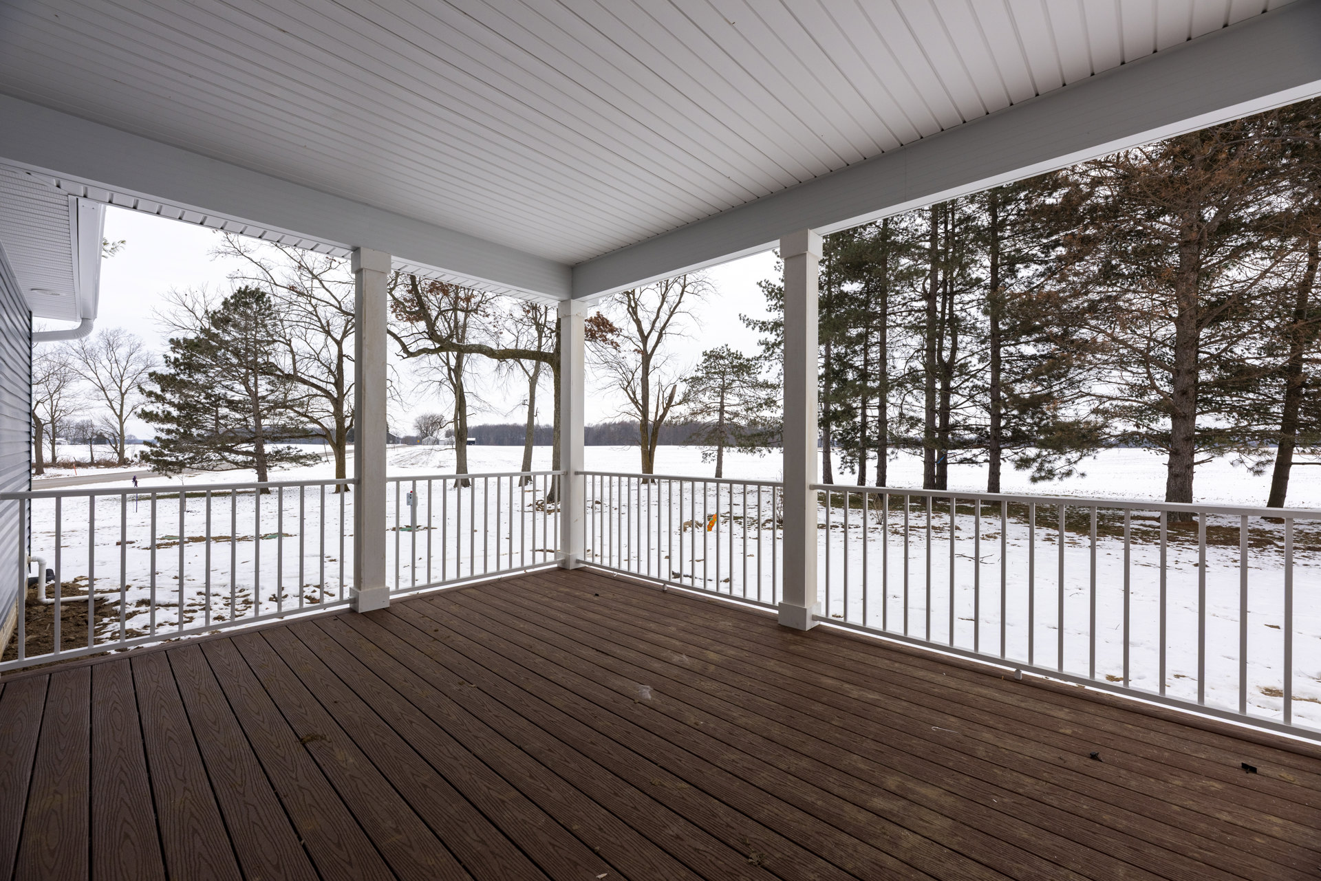 Wood deck with white railing and tall white pillar, overlooking snowy landscape with trees; white ceiling features a window, deck floor shows a few cracks.