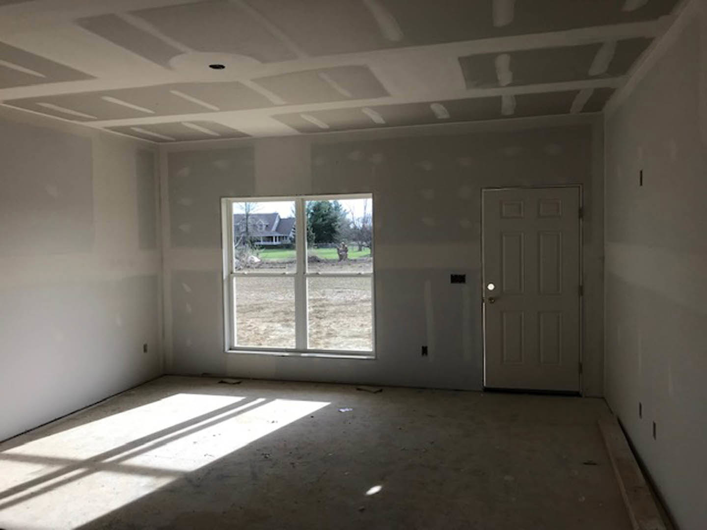 Sunlit room featuring a white square-patterned ceiling, light wood flooring, a paneled door, and a window overlooking a neighboring house.