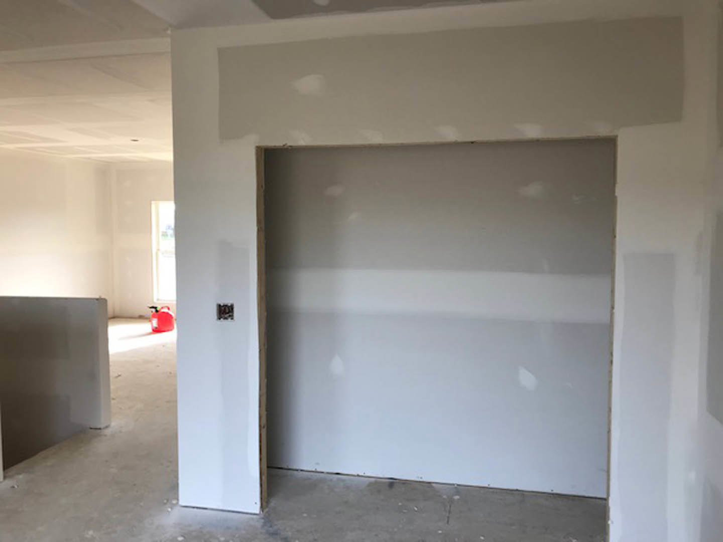 White plaster walls and ceiling, hardwood flooring, open doorway with sunlight streaming through, red gas can with black handle near the entrance.
