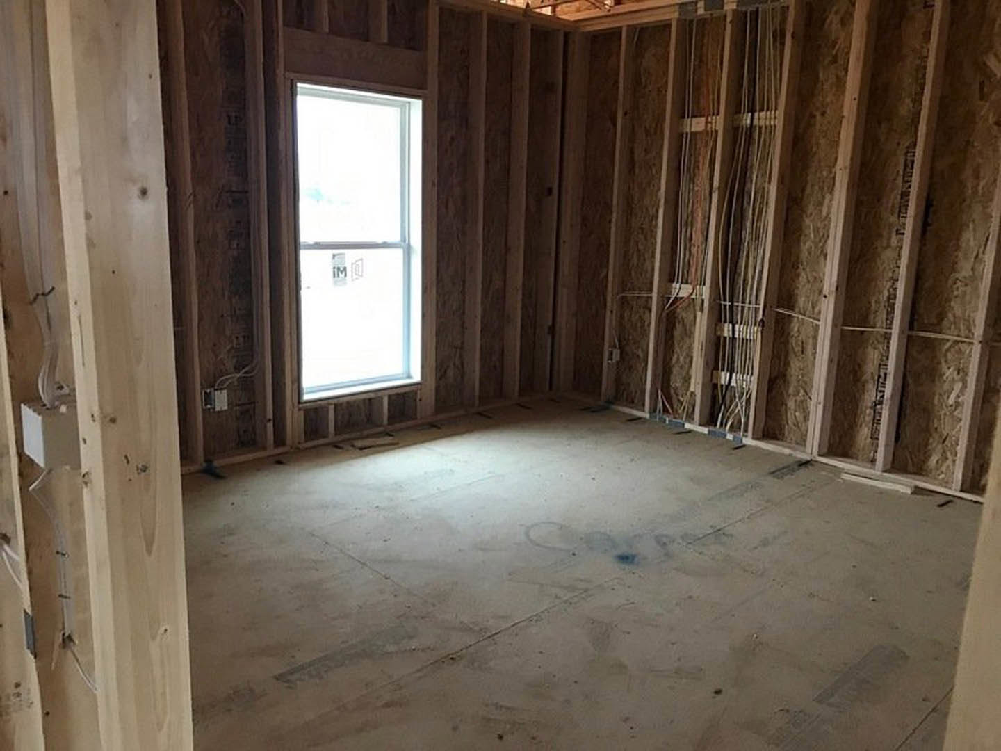 Unfinished room with exposed wooden beams, plaster walls, concrete floor, window with sign, visible electrical wires, and white utility box with attached pipe.