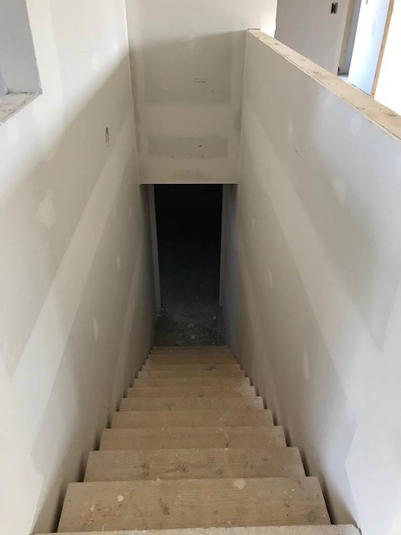 Wood staircase with open risers ascending toward a white plaster wall, door open to a dimly lit room, composite flooring visible.