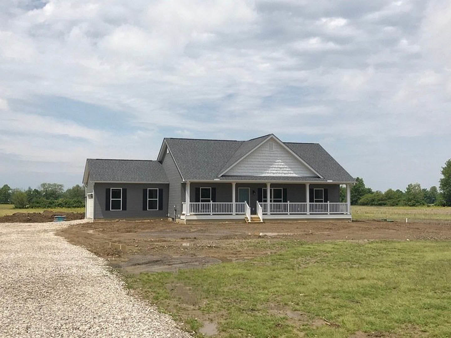 Two-story house with gray siding, white porch railing, attached garage, expansive green lawn, and mature trees under a partly cloudy sky