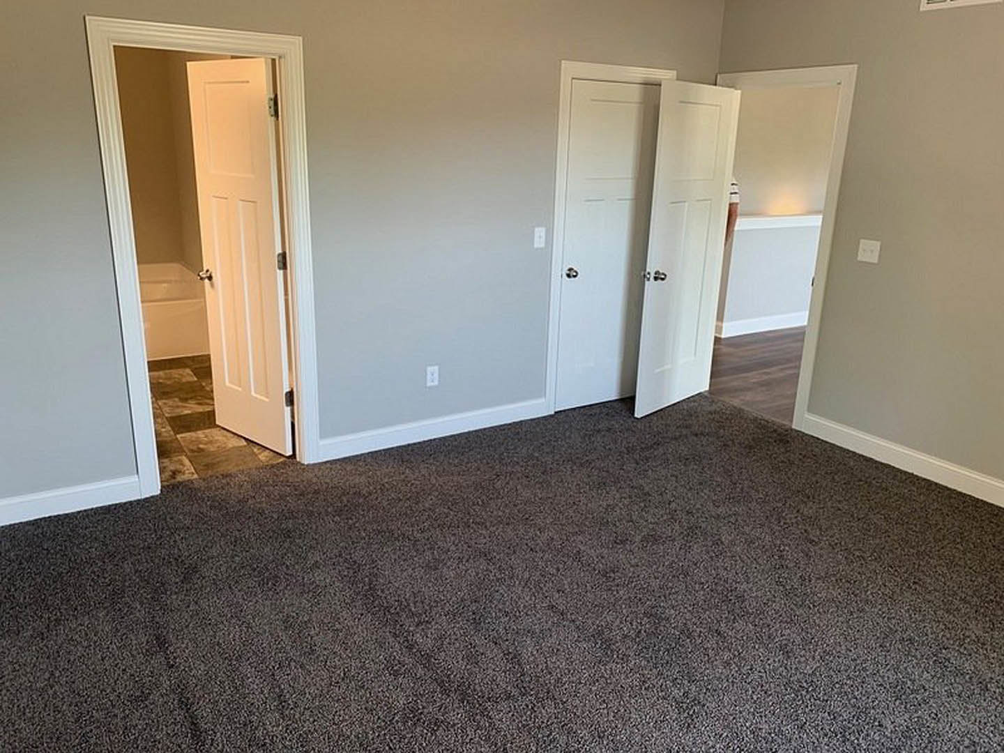 Carpeted room featuring two white doors with silver handles, white walls, and partial view of a bathroom with a bathtub