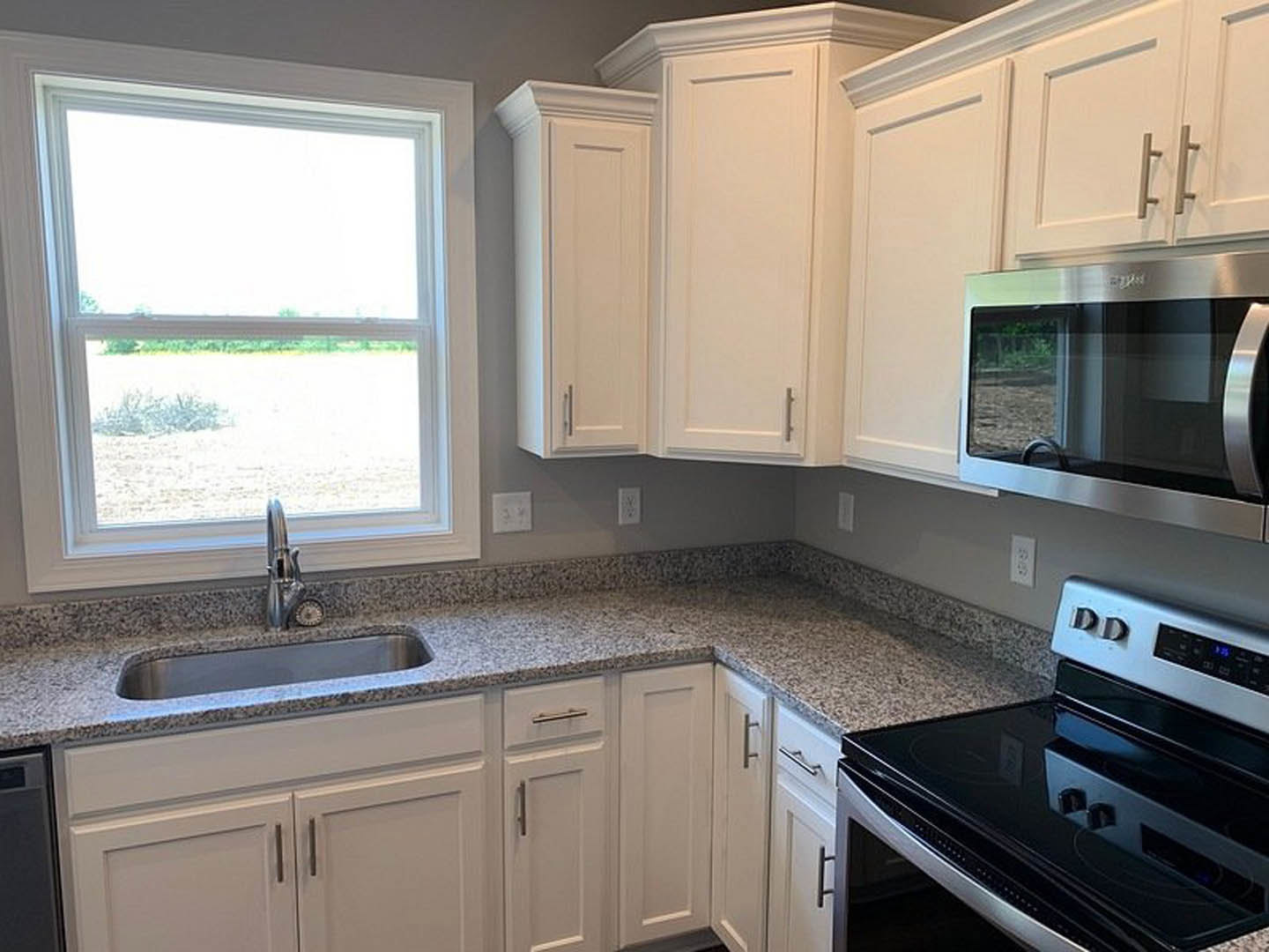 White kitchen cabinets, black stove, stainless steel microwave, grey countertop, window above sink overlooking a field, white sign with black lettering.