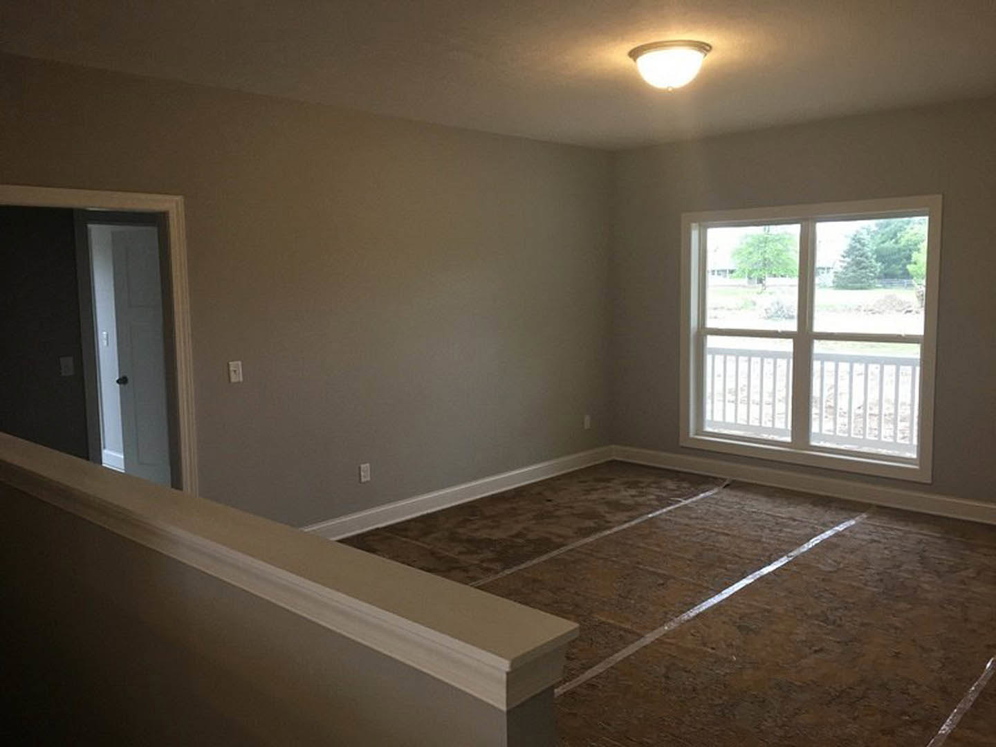Bright room with white plaster walls, dark flooring, white door, large window featuring a white railing, and ceiling light fixture turned on