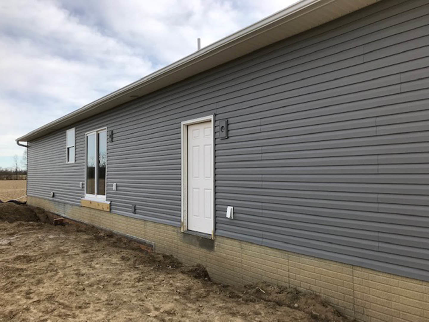 Grey siding house with white front door, black shutters, glass window, and bare dirt yard under cloudy sky