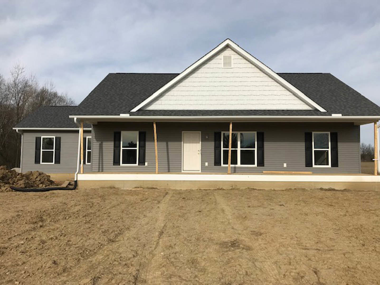 Partially built house with gray siding, black-framed white door, white-trimmed windows, dirt ground marked by tire tracks, surrounded by trees and cloudy sky