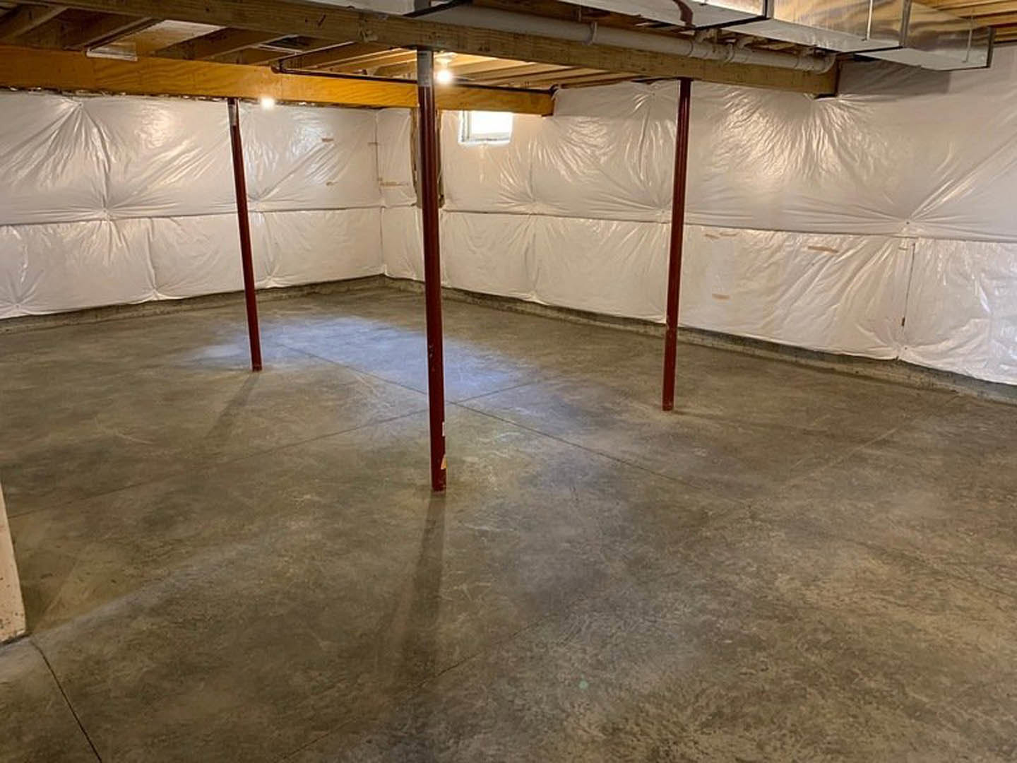 Basement with white plastic wall covering, concrete floor, red support poles, small window, and ceiling light fixture.