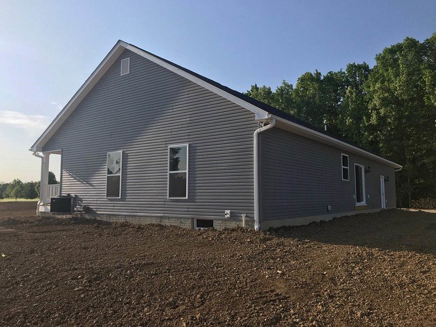 Partially built house with gray siding, white-framed square windows, shingled roof, dirt yard, and surrounding trees under a cloudy sky