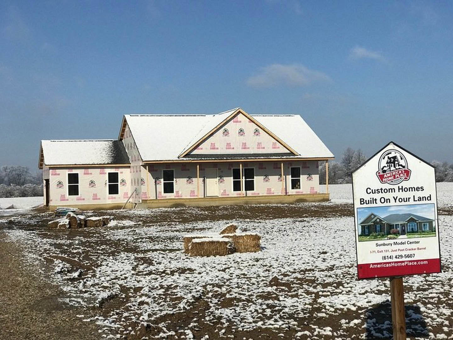 Partially built house with exposed framing and plywood, snow covering the roof and ground, construction sign visible in front, overcast winter sky in background.