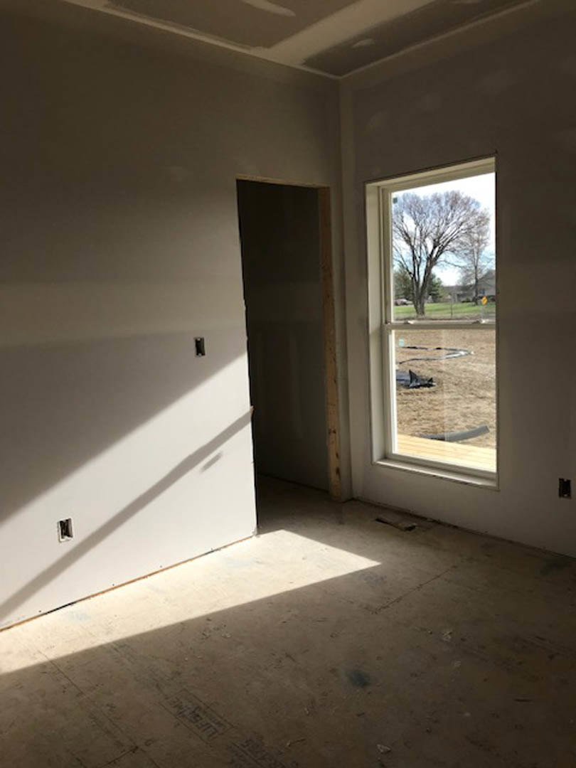Neutral-toned plaster walls, large window overlooking sandy yard and leafless tree, light wood flooring, wall-mounted light switch, sunlight streaming across floor
