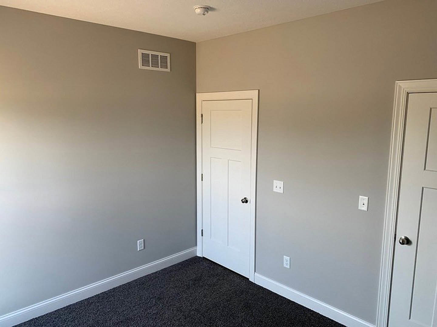 White paneled door with silver handle, wall-mounted vent, smoke detector on white ceiling, beige carpet flooring, white walls with square trim detailing