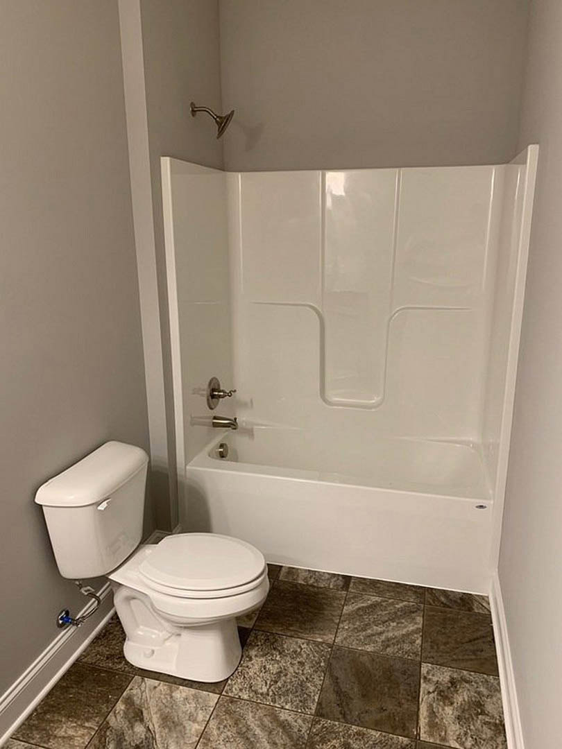 White bathtub with silver faucet beside a white toilet with closed lid and chrome handle, surrounded by light-colored tile walls and floor.