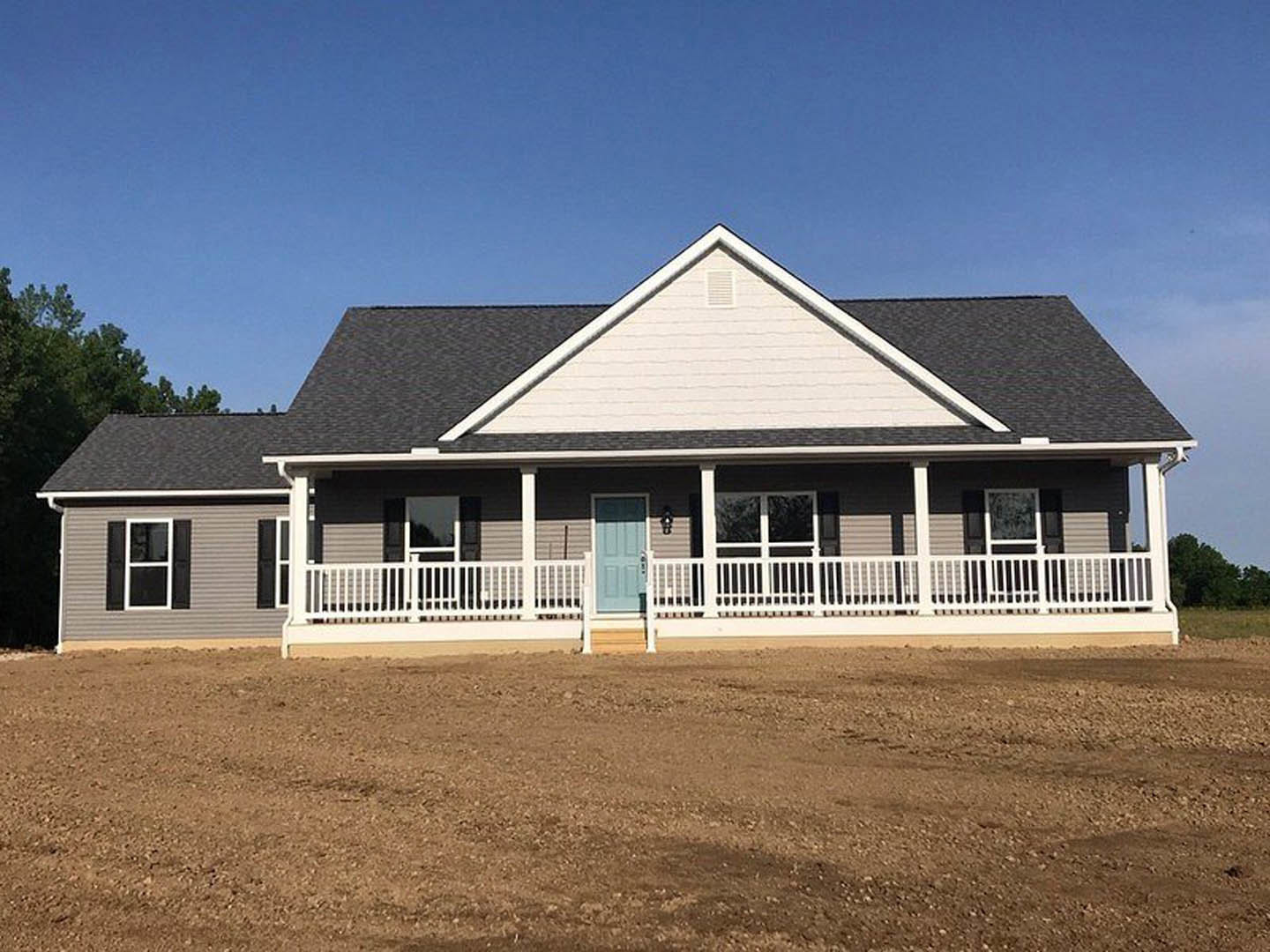 White porch with white railing, blue front door, white windows with black shutters, dirt yard, and tree in background.