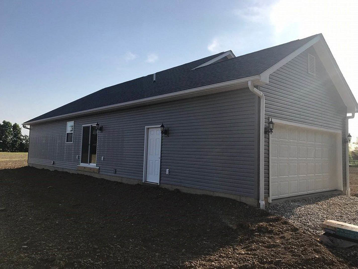 Grey siding house with black trim, white front door, white garage door with exterior lamp, large windows, surrounded by trees under partly cloudy sky