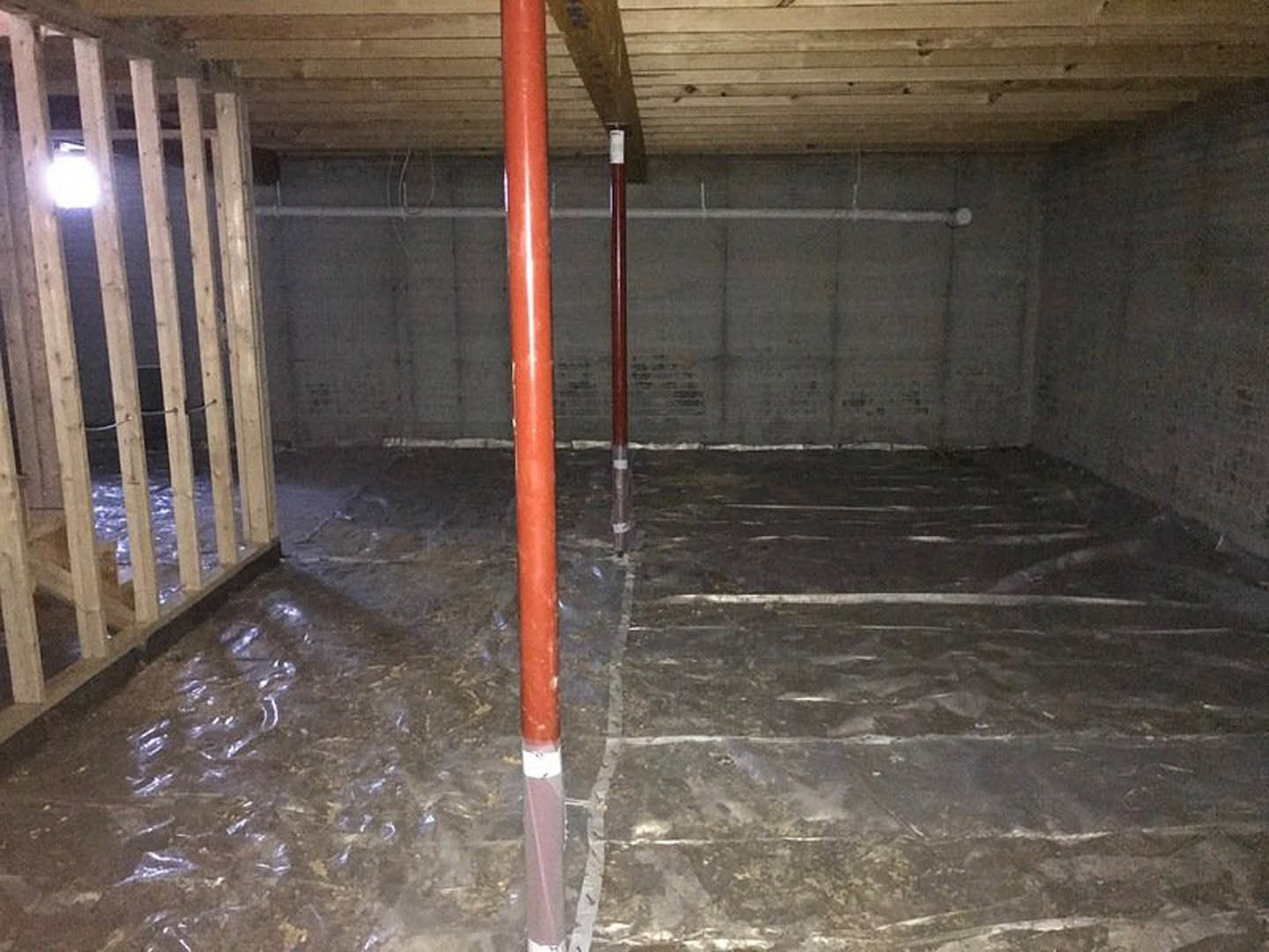 unfinished basement with exposed wooden beams, red metal support pole, light bulb mounted on wall, red pipe running along concrete floor, and insulated walls