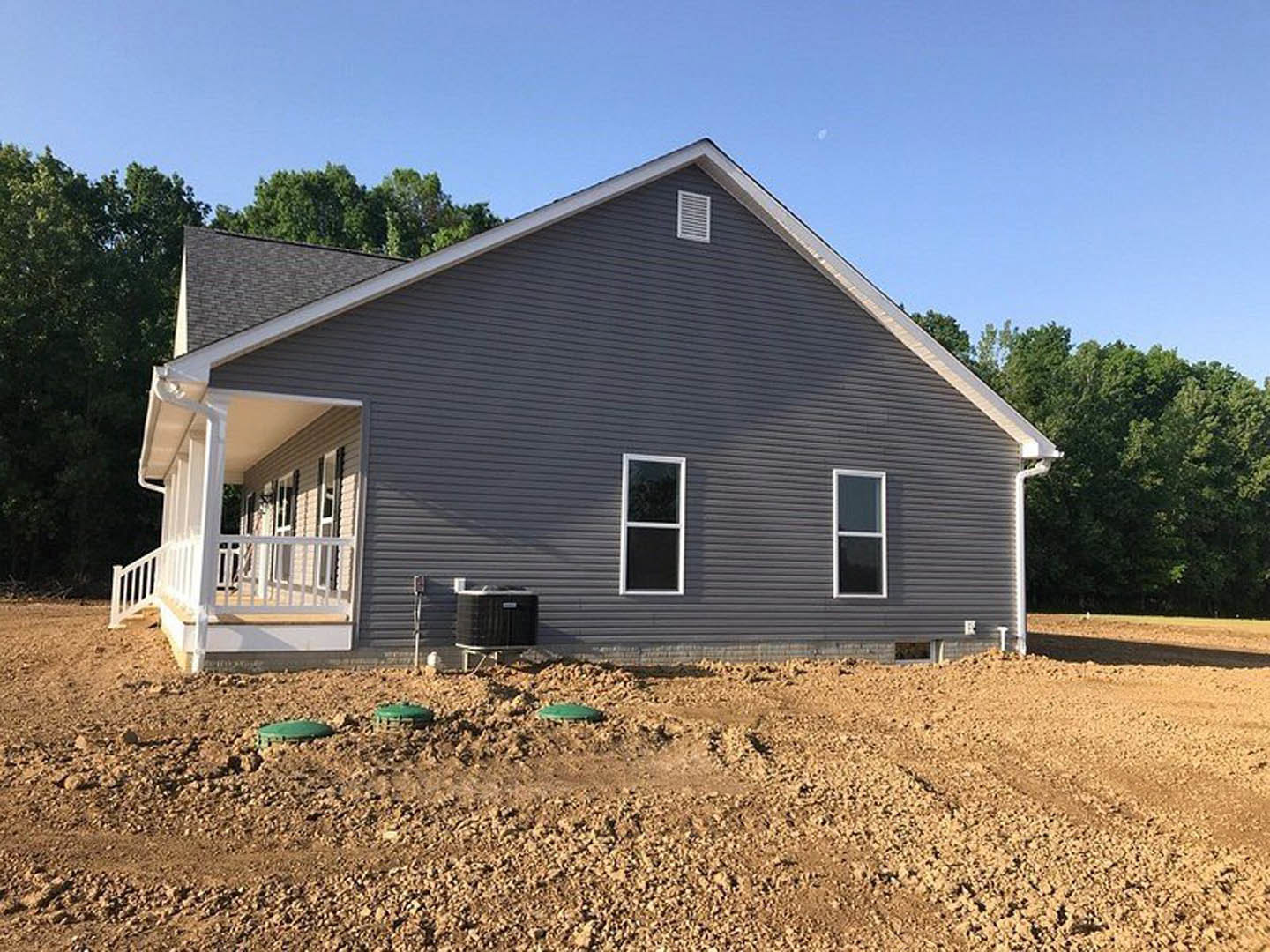 Two-story house under construction with white-framed windows, covered porch, dirt yard, green construction equipment, and mature trees in the background